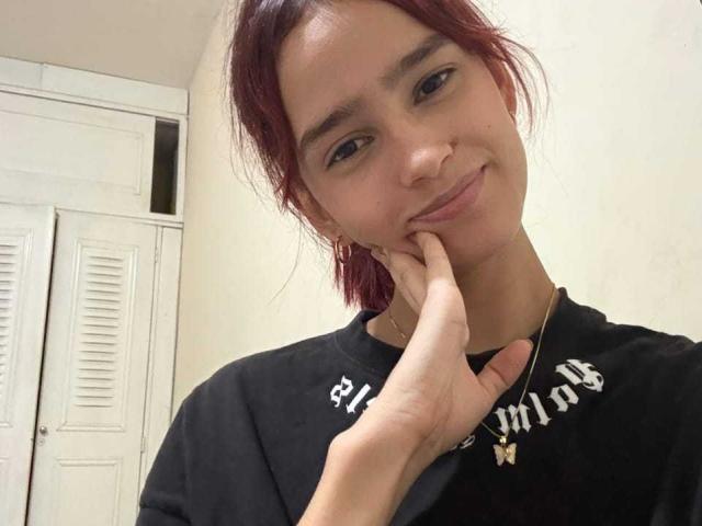 Photograph of MelodyMansson with short red hair, smiling slightly, touching her chin with one hand. Wearing a black shirt with white text. White shuttered doors in the background. Casual, slightly sexy.