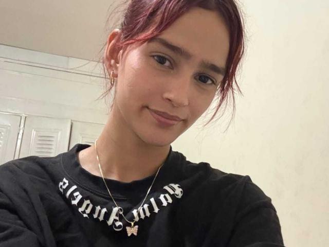 Photograph of MelodyMansson with reddish-brown hair, wearing a black "Bergman" shirt and silver necklace, smiling slightly. Background has white lockers. MelodyMansson has a subtle, sexy, relaxed vibe.