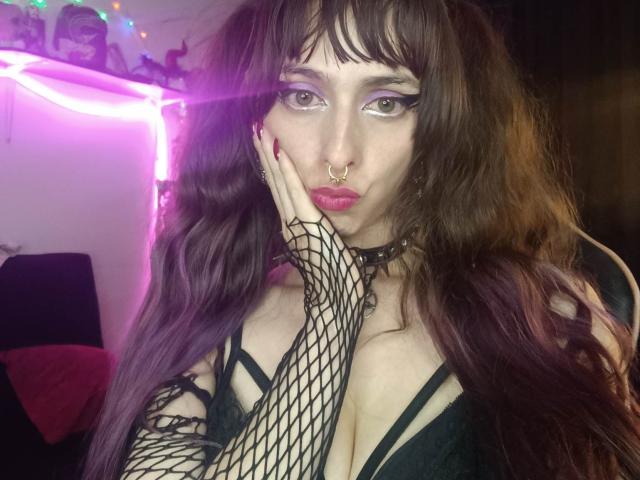 Close-up photo of KizzuKyuwu with pale skin, dark wavy hair, and heavy makeup. She's wearing a black fishnet glove, choker, and black top. Glaring seductively, hand on face, neon lights in background.