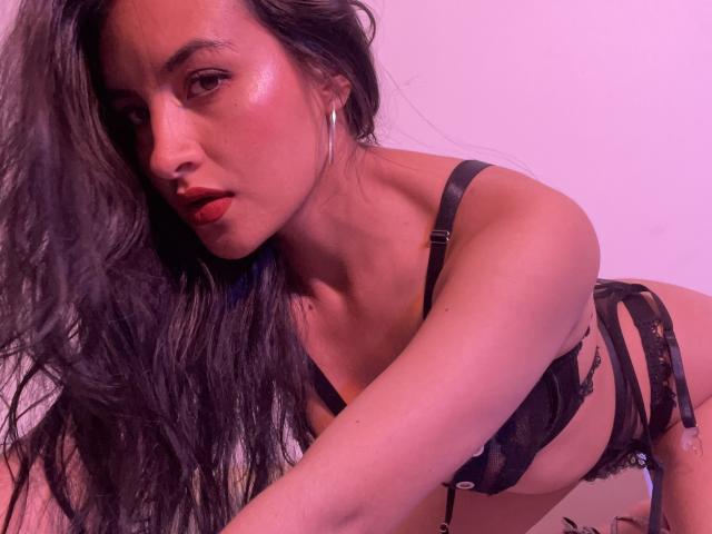 Close-up of EmilyJensenn, leaning forward with long black hair, wearing black lace lingerie, glistening skin, red lipstick, hoop earrings. Pink lighting highlights her curves, creating sexual tension.