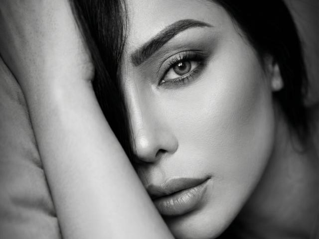 Close-up of SandraXSeins, monochrome photograph. She has half her face in shadow, intense gaze, slightly parted lips. Her skin is smooth, eyes heavily made-up. Forearm covers part of her face. Sexual tension.