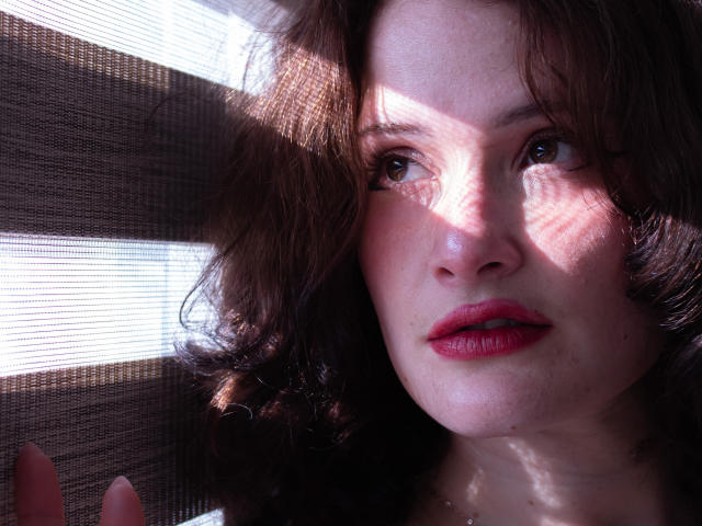 Close-up of DanaStone, a sexy woman with curled brown hair, red lips, and glowing skin. Harsh light creates intense shadows on her face. Her gaze is direct, with a seductive look.