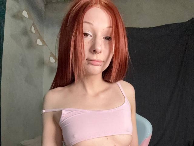 Photograph of a young woman with straight, red hair, wearing a white, off-shoulder top, light skin, and a confident expression. She has small breasts and a subtle smile. Fucking sexy.