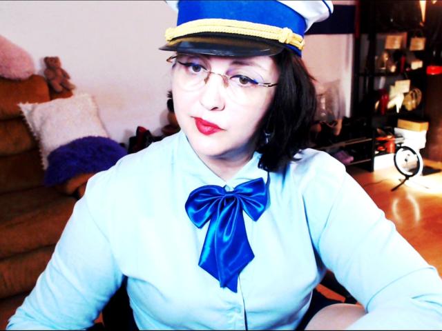 ImperatrizaSanda with pale skin and red lipstick, wearing a blue and white flight attendant outfit with a large blue bow, and a blue hat with a yellow band. Blurred indoor background.