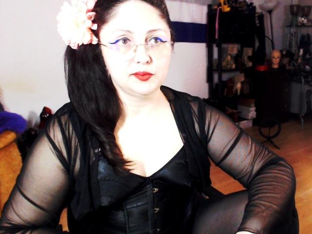 Photograph of ImperatrizaSanda, a pale-skinned woman with black hair in a ponytail, wearing a sheer black top and corset, red lipstick, bright white light on face, dark background, sexual tension.