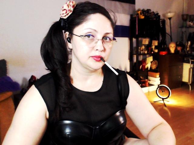 Photograph of ImperatrizaSanda, fair-skinned, black hair in a ponytail with a red patterned hair tie, wearing glasses, black leather bustier, sucking on a cigarette, indoor setting, dim light.