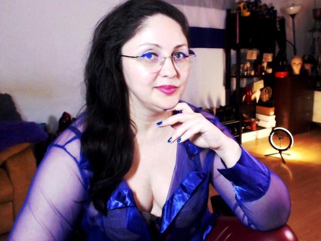 ImperatrizaSanda with pale skin, black hair, and glasses, wearing a sheer blue top that reveals cleavage. She's posing with a finger to her lips, exuding sexual tension. Bright lighting.