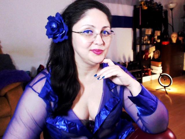 ImperatrizaSanda with pale skin and black hair, wearing a sheer blue dress with a blue rose in her hair, sitting with one hand near her face, slightly seductive expression. Bright lighting.