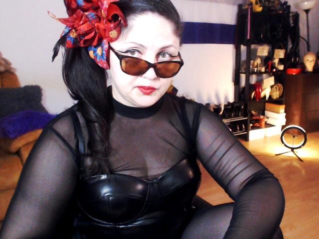 ImperatrizaSanda with pale skin and black hair, wearing a sheer black top over a black leather bustier, red flower hair accessory, brown sunglasses. Background has blurred furniture and light. Sexual tension.