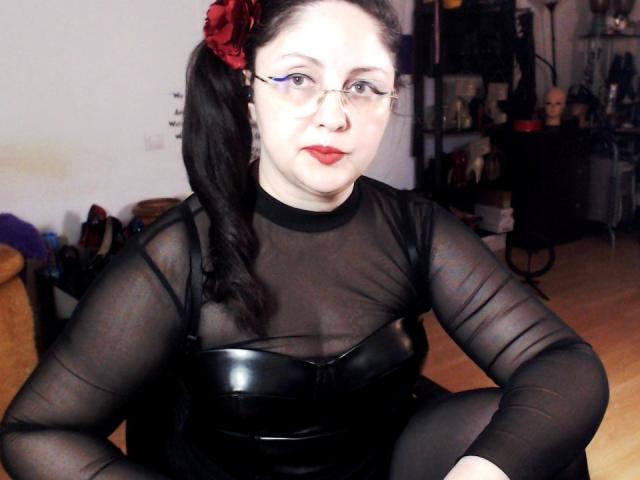 ImperatrizaSanda with pale skin and red lipstick, wearing a sheer black top and black leather bustier, black hair with a red flower, intense gaze, dimly lit room, suggestive pose.