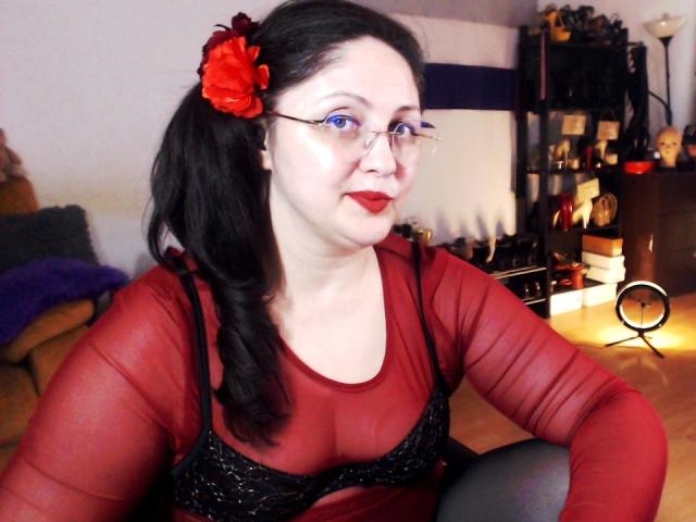 ImperatrizaSanda with pale skin and black hair, wearing red glasses and a sheer red top with a black bra, a red flower in her hair, sitting, sexy, seductive, looking to the side.