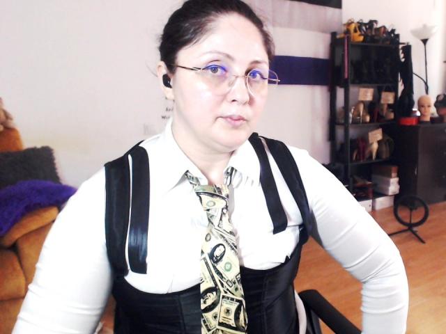 Photograph of ImperatrizaSanda with pale skin, black hair in a bun, wearing glasses, a white shirt, black corset, and patterned tie, sitting in a living room, slightly blurred, intense gaze.