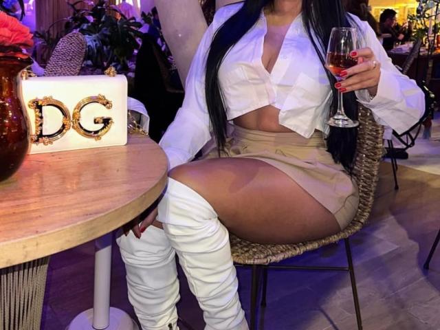 Diiana, sitting at a wooden table, wearing a white tied-up blouse revealing cleavage, beige shorts, and white thigh-high boots, holding a glass of red wine. Background: dimly lit bar.