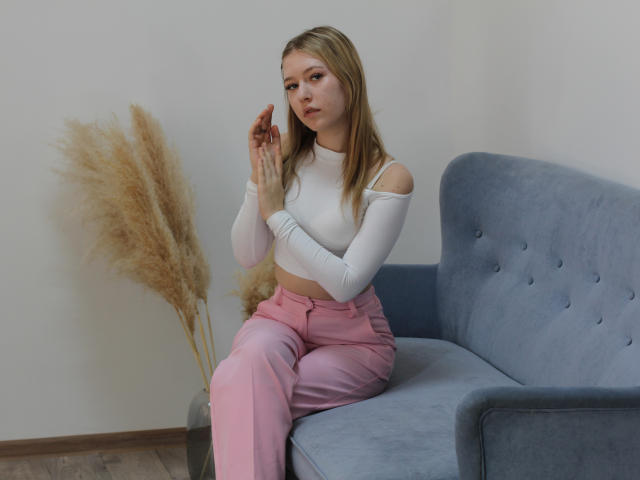 RosaVance sits on a blue sofa, wearing a white, long-sleeve crop top and pink pants. She has blonde hair, lightly touching her face with a neutral expression. Pampas grass nearby.
