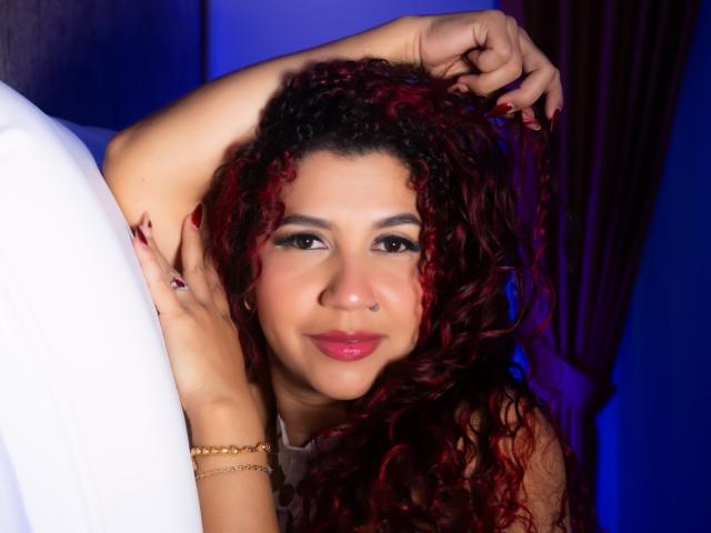 AntonelaRiveiro with curly red hair, nose ring, and red lipstick, holding her hair, smiling, wearing a gold bracelet, white shirt, and dark red curtains in the background.