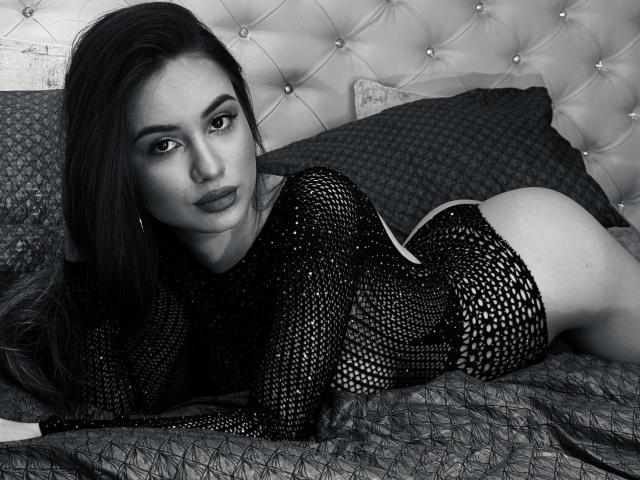 AmiellaRose, lying on a bed, wearing a black mesh top, exposing her ass, looking directly at the camera with sultry eyes, dark hair, tufted headboard, monochrome photo, sexy, provocative.