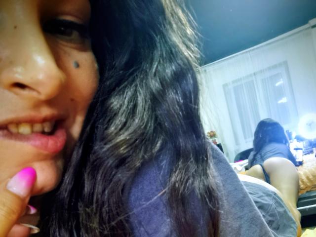 Close-up of PassionX with black wavy hair, brown skin, and a mole on her cheek, smiling with pink nail-polished finger touching her lips. Background shows another person with dark hair.