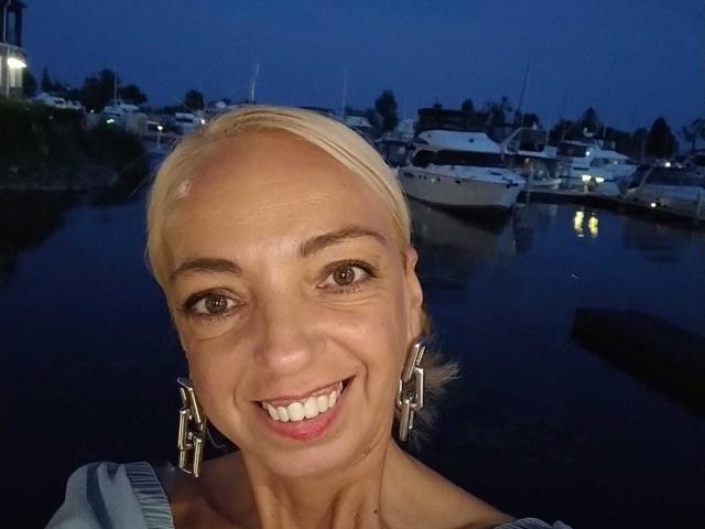 Close-up photograph of LunaBriella, a smiling woman with short blonde hair, wearing geometric earrings, against a dark blue evening marina background. Boats and lit windows in the background. Fucking sexy smile.