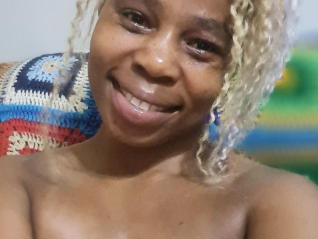 Close-up of SexyHotButt, smiling with glossy lips, dark brown skin, and curly blonde hair. Background has a colorful knit blanket and blurred green and blue hues. Focused on facial expression.