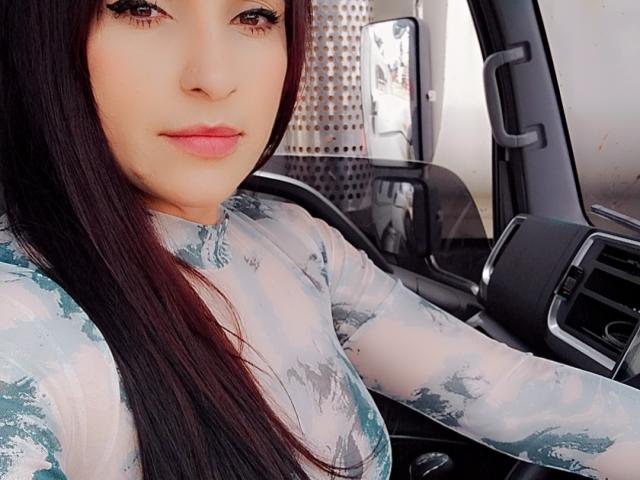 Close-up of LuluFrancaise, a woman with long black hair, wearing a white and blue patterned top, sitting in a car, hand on the steering wheel, slightly smiling, eyes focused forward.