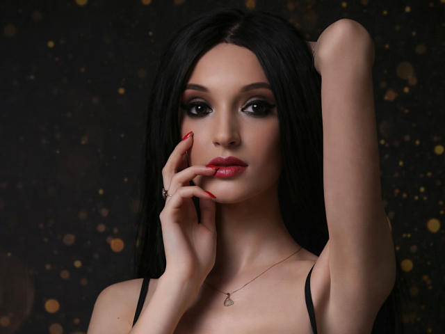 Photograph of MaiaMoxie, dark-haired woman with red lipstick, black eyeliner, and red nail polish, raising her left arm, touching her face, wearing a black strap top, dark background with bokeh lights.