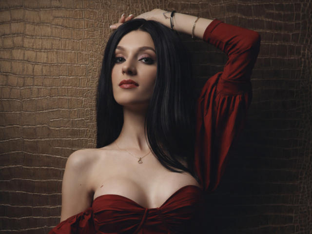 MaiaMoxie, a pale-skinned woman with long black hair, wears a red off-shoulder dress, exposing her cleavage. She's raising her right arm, hand in her hair, looking seductively at the camera.