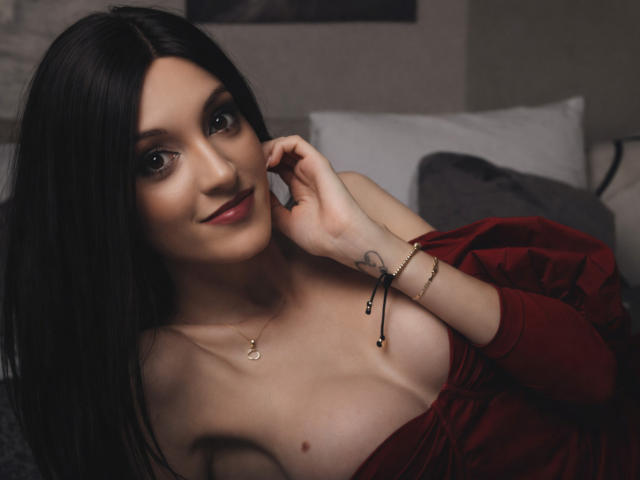 Photograph of MaiaMoxie, a slim woman with long black hair, fair skin, and small breasts, wearing a red off-shoulder top, smiling while touching her cheek, visible nipple, dark eye makeup.