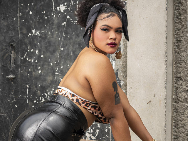 Digital image of AliceJelanni with dark skin, curly hair in a black bandana, leopard-print top, and black leather pants. She leans forward, glancing back, showing a tattoo on her right arm. Sexual tension.
