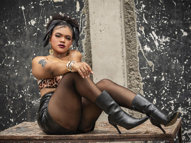 AliceJelanni, dark-skinned, with black hair, sits on a gritty ledge, legs crossed, wearing a leopard print bra, black shorts, fishnet stockings, and black boots. Tattoo on left shoulder. Fucking sexy.