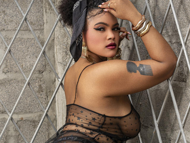 AliceJelanni with dark skin, curvy physique, wearing sheer black lingerie, black headscarf, gold earrings, bracelets, tattoo on upper arm. Leans against diamond-patterned wall, hand on head, sultry expression.