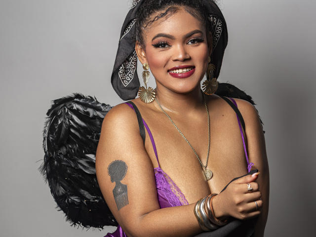 AliceJelanni with dark skin, curly hair, red lipstick, wearing a purple lace bra, black feather wings, gold jewelry, and a black headscarf. Visible tattoo on upper arm, smiling seductively.