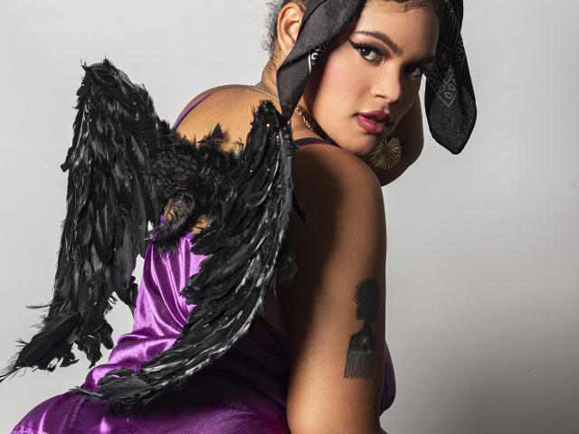 Digital artwork of AliceJelanni with black feathered wings, purple satin dress, black headscarf, and hoop earrings. She's glancing back seductively, revealing a tattoo on her shoulder. Sexual tension.