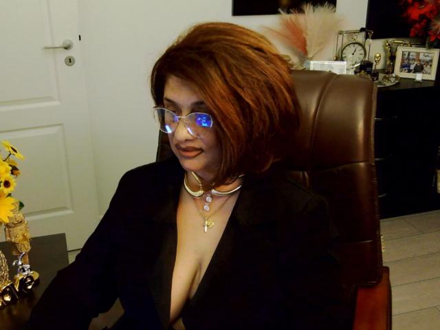 ClassybutNaughty with red hair, blue reflective glasses, deep cleavage in black suit, gold choker, and cross necklace, sitting in brown leather chair, smiling.