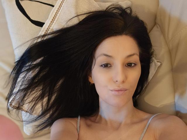 JoliDanielle lying on a pillow, black hair spread out, brown eyes, small nose, wearing a grey tank top, subtle smile, small gold earrings, soft light on her face.