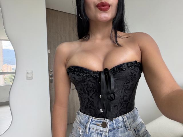 EmilyJensenn with black lace-up corset, showing her tits, wearing denim jeans, pouty red lips, black hair, and hoop earrings, in a modern white room.