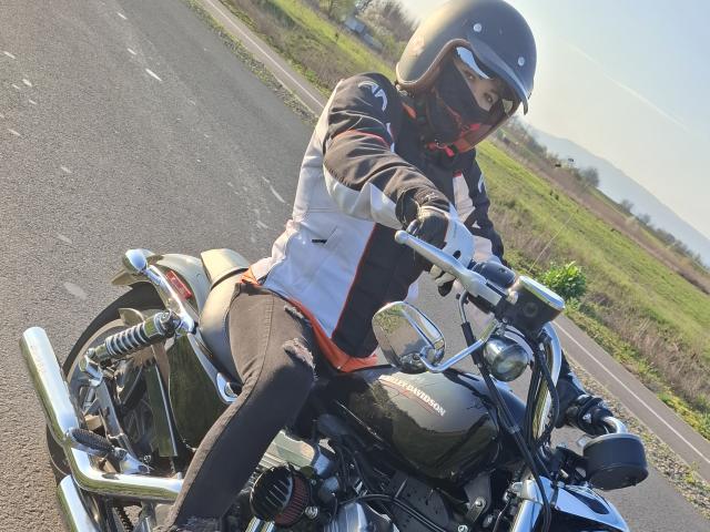 FrancaiseAnnaDom riding a motorcycle, wearing a helmet, black gloves, and a white jacket with red accents, on a road with grass and mountains in the background.