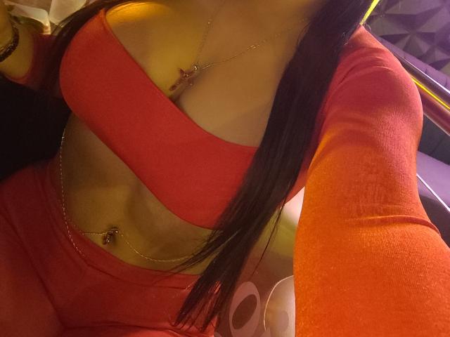 Diiana's red outfit with cutouts reveals her tits and a navel piercing. She has long brown hair and wears a necklace with a small pendant. Her abs are visible.