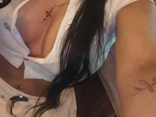 Diiana's white shirt is open, showing her tits with a red cross tattoo above. She has long black hair, a belly button piercing, and an arm tattoo with a knot.