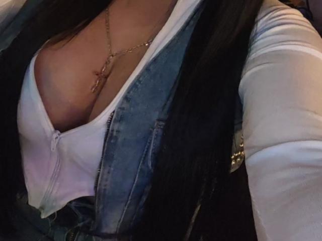 Diiana's tits are visible in a low-cut white shirt under a blue denim vest, with a gold cross necklace, in a dimly lit car.