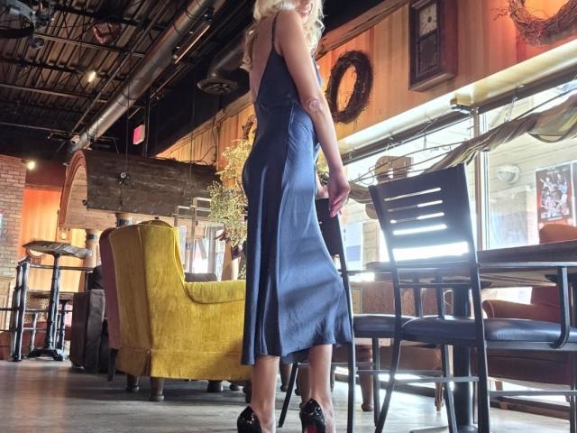 LunaBriella in a blue, silky, sleeveless dress, standing with her back to the camera, holding a black chair, wearing black heels, with wavy blonde hair.