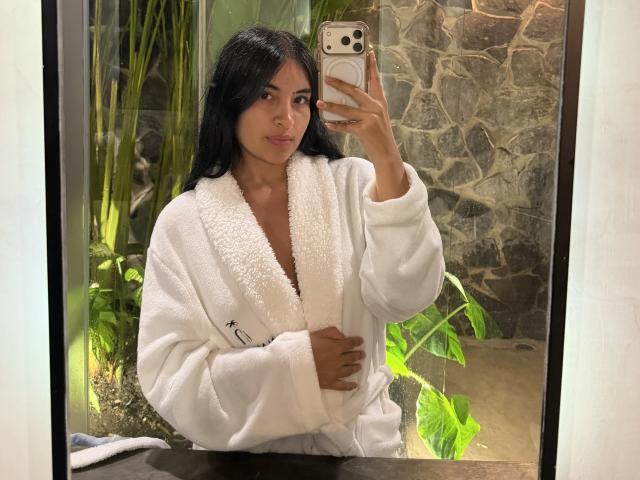 EmilyJensenn with black hair, wearing a white fluffy robe, holding a phone in front of a mirror, showing a subtle smile, with her hand on her chest.