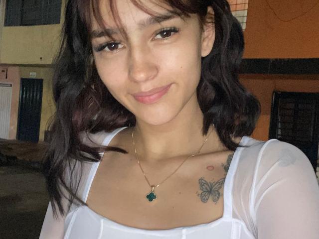 MelodyMansson with dark wavy hair, white sheer top, small green pendant, butterfly tattoos on chest, smiling, brown eyes, light skin, slight pink lip.