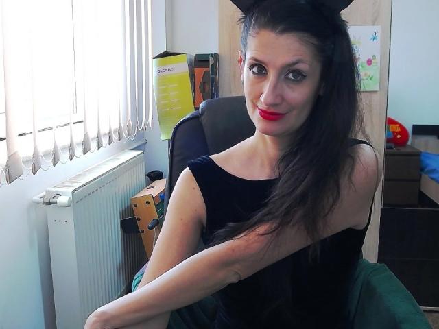 BeatrixCharmm with black cat ears, long black hair, red lipstick, black sleeveless top, sitting in a chair, green blanket, white blinds, and a radiator in the background.