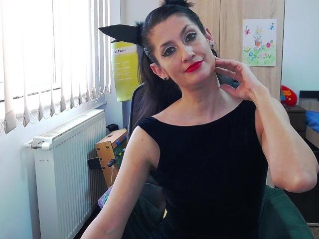 BeatrixCharmm with black cat ears, red lipstick, black sleeveless top, and hand on cheek, sitting in chair, smiling, window and radiator in background.