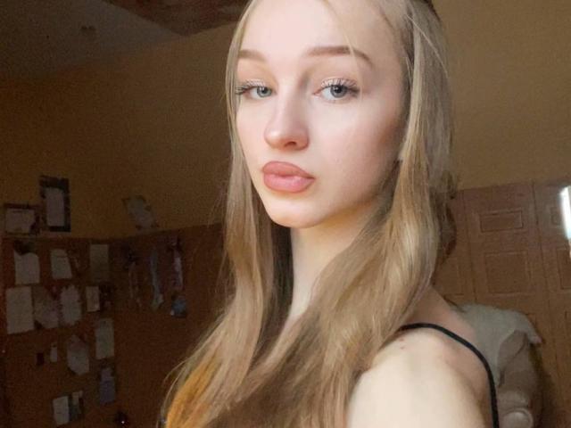 ArielleFiorello has long blonde hair, blue eyes, and pink lips. She's wearing a black strap, looking directly at the camera with a slightly pouty expression.