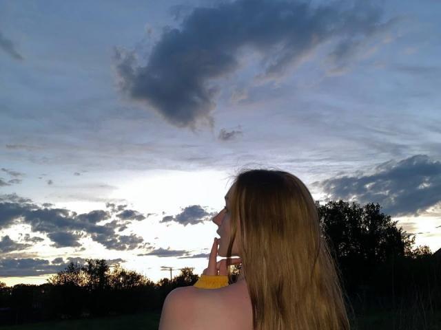 ArielleFiorello with long blonde hair, topless, holding a yellow object near her mouth, back to the camera, under a cloudy sunset sky.