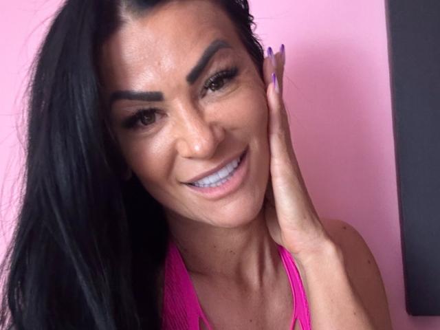 AndraD with black hair, brown eyes, and a bright smile, touching her cheek with purple nails, wearing a pink top, showing her teeth and dark eyebrows.