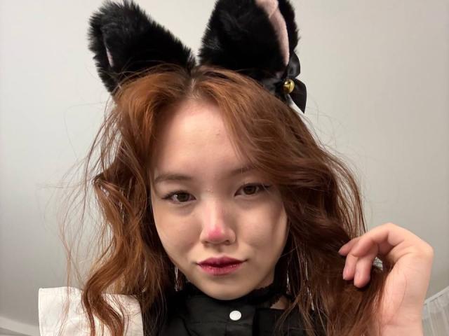 NereidaBottino with cat ears, wavy brown hair, light skin, pink nose, and red lips, wearing a black choker, looking at the camera with a slight smile.