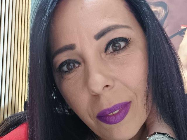 LauraSin's face with dark black hair, purple lipstick, and thick black eyeliner, showing a slight smirk, her brown eyes looking forward, wearing a red top.