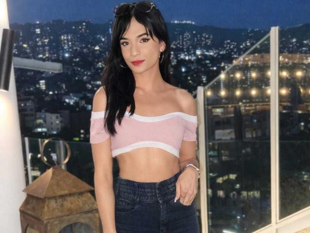 PamelaKander with black hair, red lipstick, off-shoulder pink top, showing tits, midriff, and dark jeans, standing by a glass balcony at night with city lights.